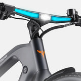 Close-up of a bicycle handlebar area with a black frame, bright blue illuminated light bars, a white center light, and an orange logo on the downtube against a light gray background.